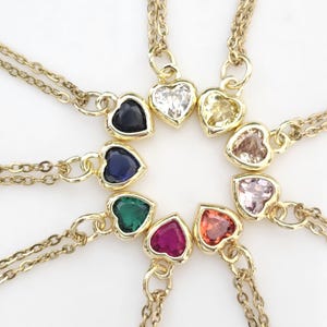 May include: A collection of gold-colored necklaces, each featuring a heart-shaped pendant with a different colored gemstone. The gemstones include clear, yellow, pink, orange, red, green, blue, and dark blue. The necklaces are arranged in a circular pattern.