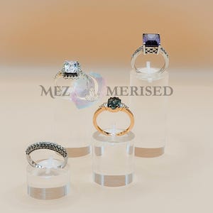Peut inclure: Plusieurs bagues présentées sur des présentoirs transparents. Les bagues présentent divers designs, dont une pierre violette taillée carrée, un anneau avec de petites pierres et une bague en or avec une pierre ovale. Les bagues sont présentées sur un fond neutre.