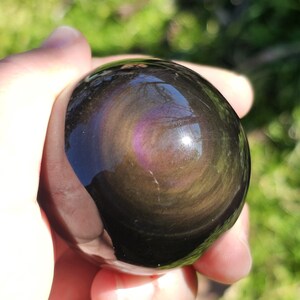 May include: A polished obsidian sphere, held in a hand, shows a swirling pattern of dark brown, gold, and purple tones. The surface reflects light, giving a glossy look. The gemstone is a natural, decorative item.