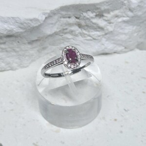 May include: A silver-toned ring featuring a central oval-shaped dark pink gemstone surrounded by a halo of small clear stones. The band is slender and includes a row of tiny clear stones embedded along the sides of the metal shank.