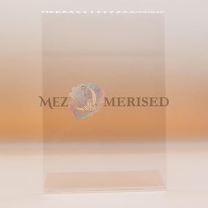 Pode incluir: Um saco de celofane transparente e retangular com as palavras "MEZ MERISED" impressas em uma fonte marrom escura. Um logotipo colorido com uma lua crescente, cristais e detalhes em aquarela está centrado no saco.