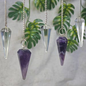 May include: Five faceted crystal pendulums hanging from silver-toned metal chains. Three pendulums are made of clear, transparent quartz, while two are made of deep purple amethyst. Each stone is cut into a pointed, multi-sided geometric shape.