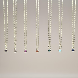 May include: Several silver necklaces, each featuring a fine chain and a small, coloured gemstone pendant. The gemstones are in shades of purple, grey, red, teal, light blue, and dark blue. The necklaces are presented against a plain backdrop.