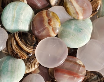 May include: A collection of polished stones in various colors and patterns. The stones include shades of brown, pink, green, and white, with visible veining and marbling. The shapes are mostly oval and round, with a smooth, glossy finish.