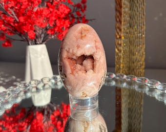 Puede incluir: Un huevo de cristal rosa y blanco con una geoda en su interior. El huevo está sobre un soporte transparente y se refleja en un espejo.