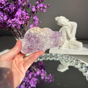 May include: A cloud-shaped amethyst crystal, displaying a light purple colour and textured surface. The image includes purple flowers and a white statue in the background. The crystal is held in a hand.