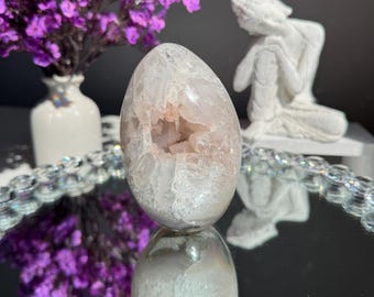 Puede incluir: Una geoda de ágata blanca y rosa con una gran abertura, que se asemeja a una forma de huevo. La geoda está sentada sobre una superficie de espejo con un arreglo de flores moradas en el fondo.