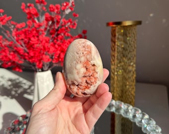 Puede incluir: Un cristal en forma de huevo de color rosa y blanco con una superficie rugosa y texturizada. El cristal tiene una base blanca con cristales rosas y rojos incrustados.
