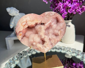 May include: A pink heart-shaped crystal geode on a gold stand. The geode has a rough, textured surface with a cavity in the center. The stand is on a mirrored surface with a decorative crystal border. Purple flowers are in the background.