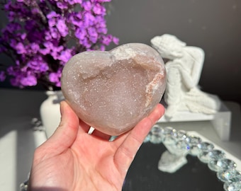 May include: A large, heart-shaped, pink and white agate geode with a crystal-lined interior. The geode is being held in a hand.
