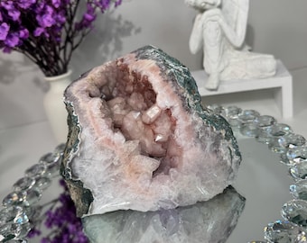 May include: A geode with a pink crystal interior and a dark green and white exterior sits on a mirrored tray with a crystal border. Purple flowers in a white vase and a white statue are in the background.