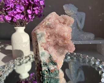 May include: A pink and green crystal cluster with a rough, natural texture. The crystal is sitting on a mirrored surface with a decorative border.
