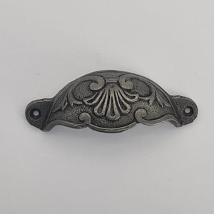May include: An ornate drawer pull in a dark grey, antique finish. The metal handle features a curved shape with a central fan design and two holes for mounting. The detailed pattern adds a vintage aesthetic.