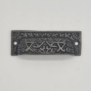 May include: A silver-toned metal drawer pull with an ornate design. The pull has a rectangular shape with a curved top and bottom. The design features a repeating pattern of stylised leaves and flowers.