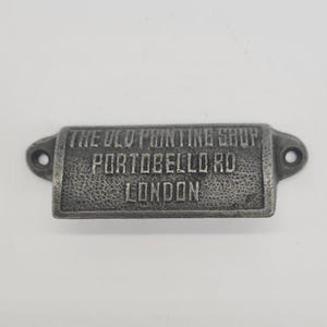 May include: A rectangular, antique-style metal drawer pull with the text "THE OLD PRINTING SHOP PORTABELLO RD LONDON" embossed on its surface. The pull has a textured, aged appearance and features two holes for mounting.