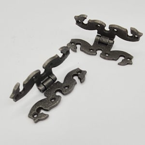 May include: Two antique-style hinges with a decorative, ornate design. The hinges are made of metal and have a dark grey finish.