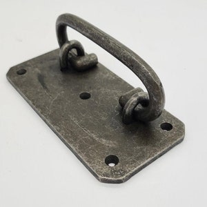 May include: A rustic, hand-forged iron door handle with a rectangular base and a curved handle. The handle is finished in a dark grey colour and has a distressed look.