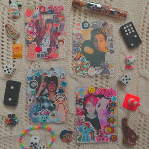 Pode incluir: Uma variedade de itens decorativos, incluindo quatro molduras com fotos de pessoas, rodeadas por adesivos e enfeites. Outros itens incluem dados, um controle remoto, uma pulseira e pequenas figuras, todos dispostos em uma superfície texturizada de cor creme.