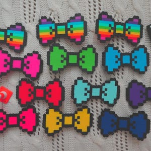 Puede incluir: Una colección de pajaritas pixeladas de colores, incluyendo arcoíris, rosa, verde, azul, rojo, amarillo y morado. Las pajaritas están dispuestas sobre una superficie blanca texturizada, con algunos pequeños accesorios dispersos.