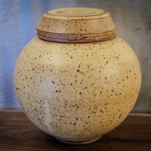 May include: A speckled, round ceramic jar with a matching lid. The jar is a light beige color with brown speckles, and the lid has a circular design on top. The jar is a decorative item.