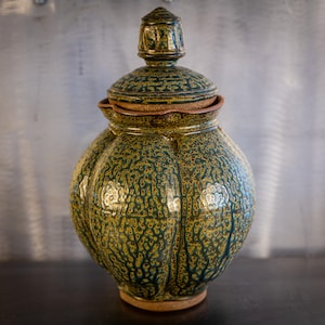 May include: A green and brown ceramic jar with a lid. The jar has a textured surface and a rounded shape. The lid has a pointed top.