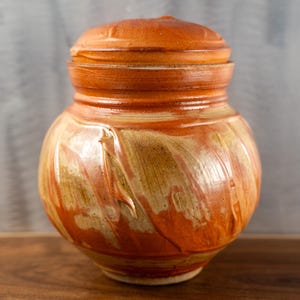 May include: A handcrafted ceramic jar with a lid. The jar is a warm orange color with streaks of tan and cream. The lid fits snugly on top. The jar is likely used for storage and is a decorative item.
