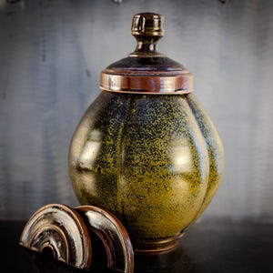 May include: A ceramic jar with a lid, featuring a dark green and brown speckled glaze. The jar has a rounded, bulbous shape and a slightly flared opening. The lid has a simple, rounded knob.