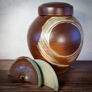May include: A brown ceramic jar with a lid and a decorative white swirl design. Two matching ceramic lid pieces are in front of the jar. The jar is on a wooden surface.