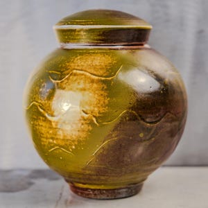 May include: A handcrafted ceramic urn with a lid. The urn is a rounded shape, with a glossy finish in shades of olive green, brown, and gold. The surface features a textured, wave-like design.