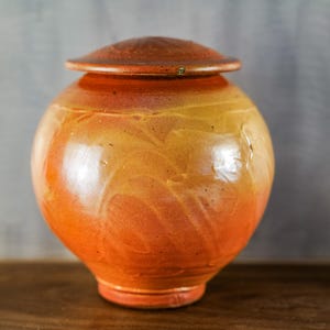May include: A ceramic urn with a lid. The urn is a warm blend of orange and yellow hues, with a glossy finish. The lid is a darker shade of brown. The urn has a rounded body and a slightly flared base. The overall height is approximately 8 inches.