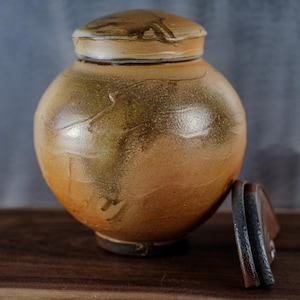 May include: A handcrafted ceramic urn with a lid, featuring a warm, earthy color palette of browns and tans. The urn has a rounded body and a lid with a similar finish. The lid is slightly ajar, revealing the interior. The urn is a decorative piece.
