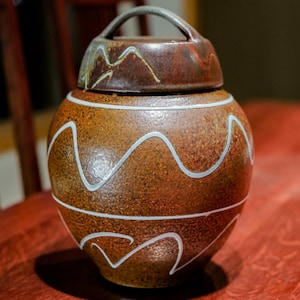 May include: A brown ceramic jar with a lid and a handle. The jar has white wavy lines painted on it.