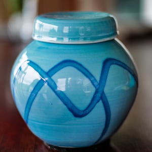 May include: A small, round, ceramic jar with a lid. The jar is a light turquoise color with darker blue, intersecting, wavy lines. The lid is the same turquoise color as the jar. The jar is sitting on a dark brown surface.