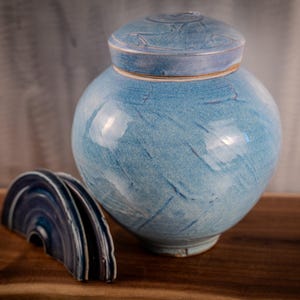 May include: A blue ceramic urn with a matching lid, featuring a round body and a textured surface. The lid is off to the side, revealing the interior. The urn is on a wooden surface.