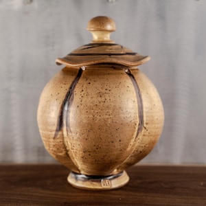 May include: A handcrafted ceramic jar with a lid. The jar is a light brown color with dark brown vertical stripes and speckles. The lid has a knob and a striped design. The jar sits on a small base.