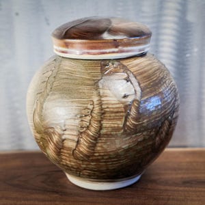 May include: A ceramic jar with a lid. The jar is round with a brown and tan marbled design. The lid is a darker brown with a tan rim. The jar sits on a wooden surface.
