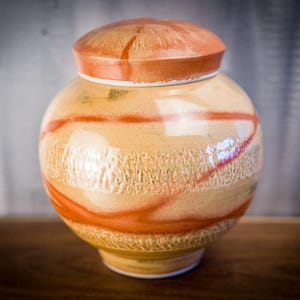 May include: A ceramic urn with a lid. The urn is a light beige color with textured vertical lines and a horizontal orange stripe. The lid is orange with a similar textured design. The urn is sitting on a wooden surface.