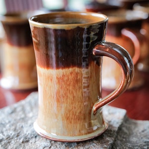 May include: A brown and tan ceramic mug with a wide base and a handle. The mug has a glazed finish and a rustic look.