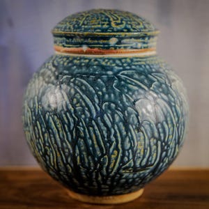 May include: A ceramic jar with a lid, featuring a textured, glazed surface in shades of teal and yellow. The jar has a round body and a decorative lid with a detailed pattern. The base is a light tan color.