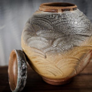 May include: A handcrafted ceramic jar with a lid, featuring a textured surface with a wave-like pattern. The jar has a color gradient from light brown to gray, with a darker brown rim. The lid is placed beside the jar.