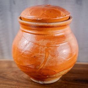 May include: A handmade, orange ceramic jar with a lid. The jar has a rounded body and a lid that fits snugly on top. The surface has a textured appearance with visible brushstrokes. The jar is likely used for storage.