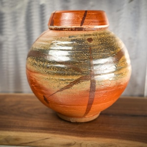 May include: A round ceramic vase with a wide body and a narrow neck. The surface features a blend of earthy orange, brown, and dark charcoal tones with horizontal ridges and a distinct dark brown line pattern wrapping around the exterior.