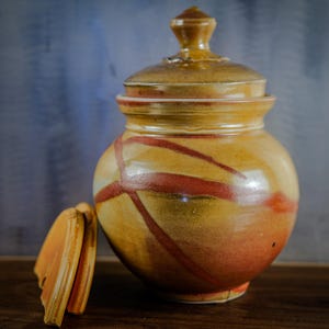 May include: A handcrafted ceramic jar with a lid, featuring a warm color palette of yellow, orange, and brown. The jar has a rounded shape with a lid and is decorated with red diagonal lines. The lid is off to the side.