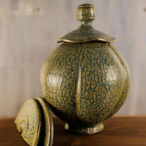 May include: A ceramic jar with a lid, featuring a textured surface with a mottled pattern in shades of teal, yellow, and brown. The jar has a rounded shape with vertical ridges and a matching lid. The lid is partially removed, revealing the interior.