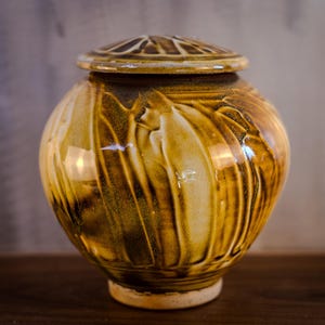 May include: A ceramic urn with a lid. The urn is a rounded shape with a glossy, textured surface. The color is a blend of golden yellow and brown, with vertical streaks. The lid has a decorative pattern. The urn is likely used for storing ashes.