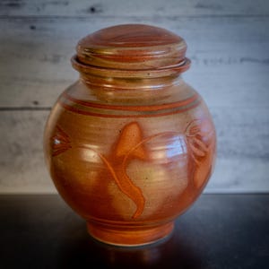 May include: A brown ceramic urn with a lid, featuring a floral design in shades of orange and brown.