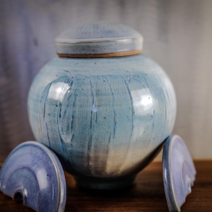 May include: A handcrafted, blue ceramic urn with a lid. The urn has a rounded shape with a light blue glaze that drips down the sides. Two broken pieces of the lid are in the foreground. The urn is on a wooden surface.