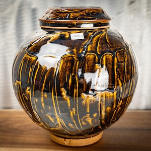 May include: A round ceramic jar with a lid, featuring a glossy dark brown and mustard yellow drip glaze pattern. The surface has vertical ridges and a small white rectangular sticker with the text 0528-10 on the upper section of the jar body.