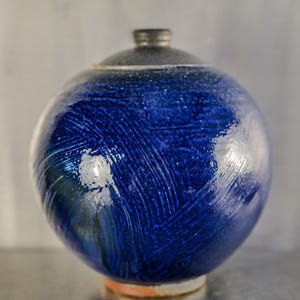 May include: A spherical ceramic vase with a glossy, deep blue glaze. The surface features textured brushstrokes and a dark gray lid. The vase is a decorative item, suitable for home decor.