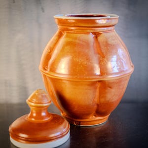 May include: A ceramic jar with a lid, both in a warm orange color. The jar has a rounded, bulbous shape with a slightly flared opening. The lid has a knob on top.